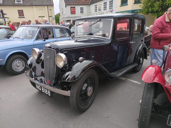A black 1934 Austin Lichfield sits in stately splendour between a more modern 1970s Triumph in blue and a red car of similar age to the Austin. The Lichfield is upright and square-edged, looking like a slightly smaller limousine. There's very little decoration to relieve its blackness, other than chrome lights, grille and small hubcaps, and a red leather interior. They're in a market square with 18th century buildings behind, and a decent crowd of people in between.