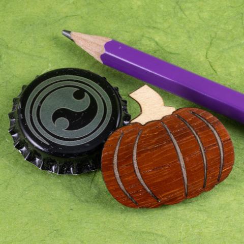 A small pin in the shape of a pumpkin made from orangey padauk with ribs of inlaid walnut and a white oak stem resting on a background of green paper along with a bottle cap and the end of a pencil to show scale.