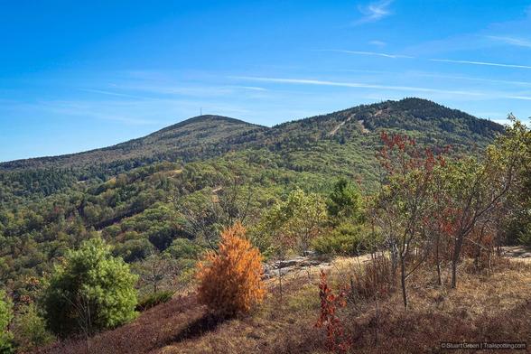 A panoramic view of a forested mountain landscape in early autumn. Two prominent peaks rise from the densely wooded terrain—one on the left crowned by a small tower or structure, the other slightly lower and more rounded. The forest displays a mix of deep green evergreens and deciduous trees transitioning to warm hues of orange, yellow, and rust. In the foreground, a rocky outcrop juts into view, dotted with sparse shrubs and young trees showing signs of seasonal change.