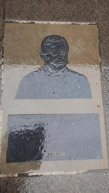 A rain covered memorial laid as a paving stone, depicting a head and shoulders relief image of a Victorian gent in spectacles, with a large moustache