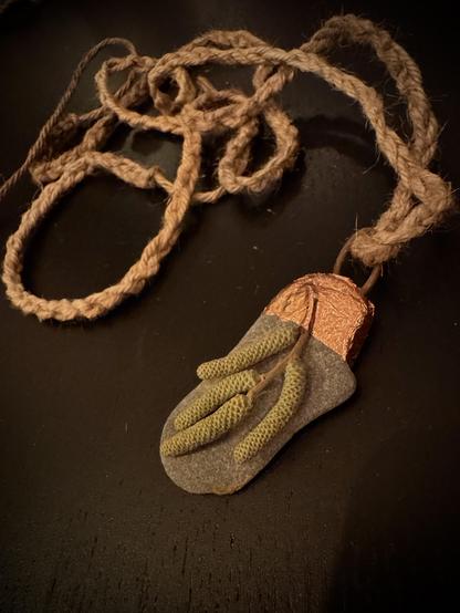 Pendant made of a catkin on a small, grey, flat rock, decorated with bronze tape and attached to a braided, natural fibre rope.