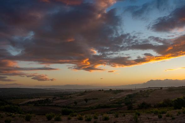 cloudy sky lit by the sunset to the right of frame, over rolling hills with the brown of summer still