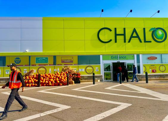 Under a vibrant blue sky, the modern exterior of "Chalo FreshCo" is showcased, featuring a striking lime green facade with the store's name and logo. The entrance is busy with shoppers entering. In front, a large, cheerful display of orange pumpkins and straw bales, along with autumnal flowers, creates a festive autumn atmosphere. Festive "Happy Diwali" banners are visible on the store windows, alongside advertisements for various food products, hinting at a multicultural shopping experience. A worker in an orange safety vest walks across the foreground, adding a sense of everyday activity to the scene.