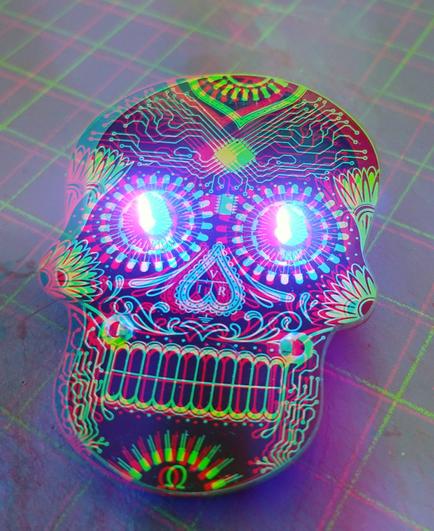 A black and gold circuit board in the style of a Day of the Dead skull, with glowing blue LEDs as eyes. Color anaglyph