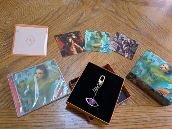 a spread of Taylor Swift Life of a Showgirl boxed set items laid out across a wooden table. Center is a pendant keychain with a purple glittered eye and a T.S. on a chain in an orange glittered and velvet case. The CD and Taylor's showgirl photos are arranged around it.