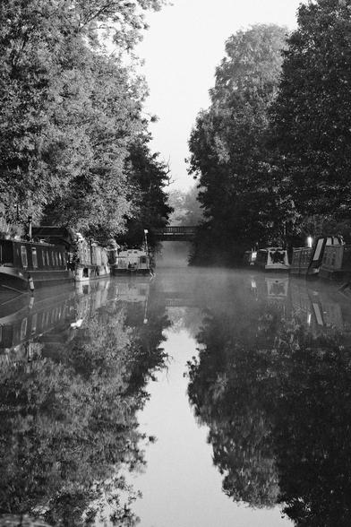 Hourglass shaped with sly and trees and sky at the top and its reflection in the canal with bridge and fog in the middle.