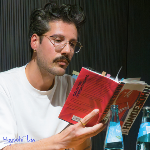 Der Autor blättert in seinem Roman "Hundedohn" um vorzulesen. Im Buch kleben mehrere bunte Marker.