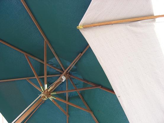 Looking up at the underside of a dark green cloth umbrella with wooden ribs. There is a partial view of the underside of a white cloth umbrella in front of the green one.