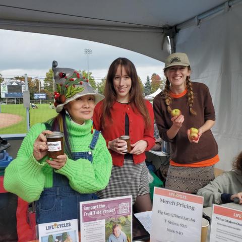 Three women standing behind a table, likely at a farmers market, promoting a "Life Cycles" fruit tree project, one woman holding a jar of fig jam, two others holding apples, with signs mentioning fruit jam, t-shirts, hats, and donation details.