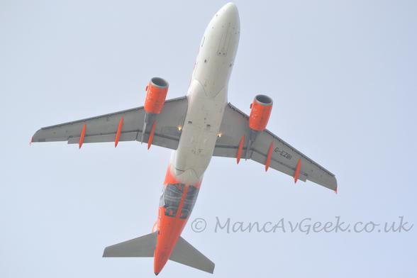 View from below o a twin engined jet airliner flying pretty much directly overhead at a very low altitude, with undercarriage tucked away, flaps extended from the rear of the wing, and the nose raised, suggesting it has just taken off.
The plane is largely white, with an orange rear fuselage overlaid with the grey head and shoulders of a historical figure.
The registration "G-EZBI" is on the lower rear fuselage in white, and under the left wing in black.
The engine pods under the wings, as well as the flap track covers, are also bright orange.
Pale grey-blue sky fills the background.