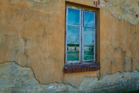Die Außenwand eines Gebäudes. Der Putz ist stellenweise bereits verschwunden. Dort wo er sich noch befindet ist die Wand ocker/gelb gestrichen. In der Bildmitte befindet sich noch ein Fenster. In ihm spiegelt sich eine der Steinbaracken.