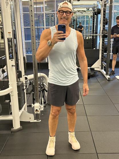 A fit individual (me) is taking a selfie in a gym. They are wearing a light tank top, grey shorts, and light-colored sneakers. The background features gym equipment and other gym-goers.
