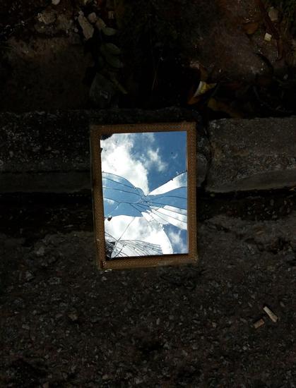Foto de um espelho retangular trincado, no chão, refletindo o céu azul com nuvens brancas.