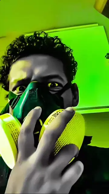 Foto com tons de verde e amarelo, um homem com cabelos curtos e cacheados utilizando uma mascara respiratória que cobre do nariz para baixo com a mão em cima de um dos dois filtros redondos da máscara