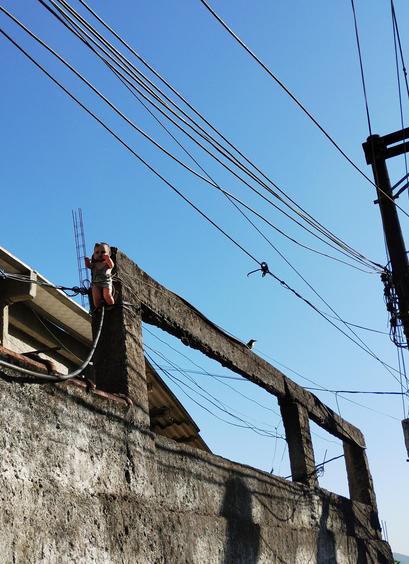 Foto de um muro de uma casa onde tem uma viga com um boneco de bebê amarrado nela. Tudo ta meio sujo e abandonado dando um ar bizarro. Tem um bem-te-vi pousado ali perto também. O céu ta bem azul lá atrás