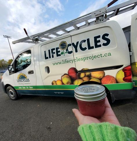 A hand in a light green sweater holds a jar of red-colored crab apple preserves in front of a white LifeCycles van with a ladder on top. The van is parked on asphalt under a blue sky with some clouds; the side of the van is decorated with a sunflower and fruit images.