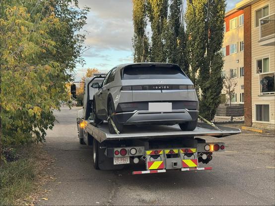 My Hyundai Ioniq 5 being transported on the back of a flatbed tow truck. The vehicle is facing away from the camera, showing its rear end and roofline with a spoiler. A license plate is visible on the tow truck's trailer, reading “23-P861”. Several orange and yellow reflective warning triangles are attached to the back of the trailer. The scene is outdoors with a building and trees visible in the background.