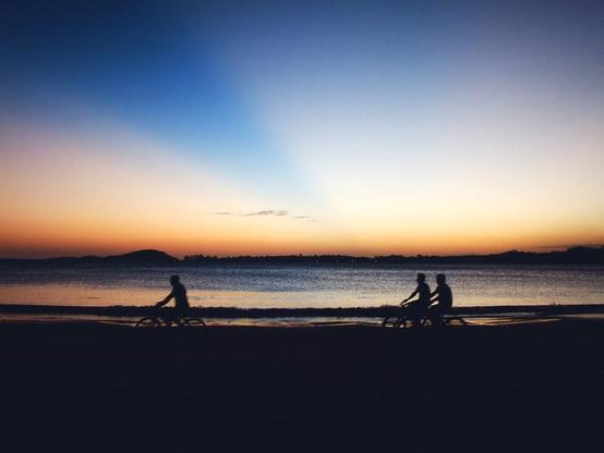 Foto de uma baía ao pôr-do-sol, com a silhueta de três ciclistas.