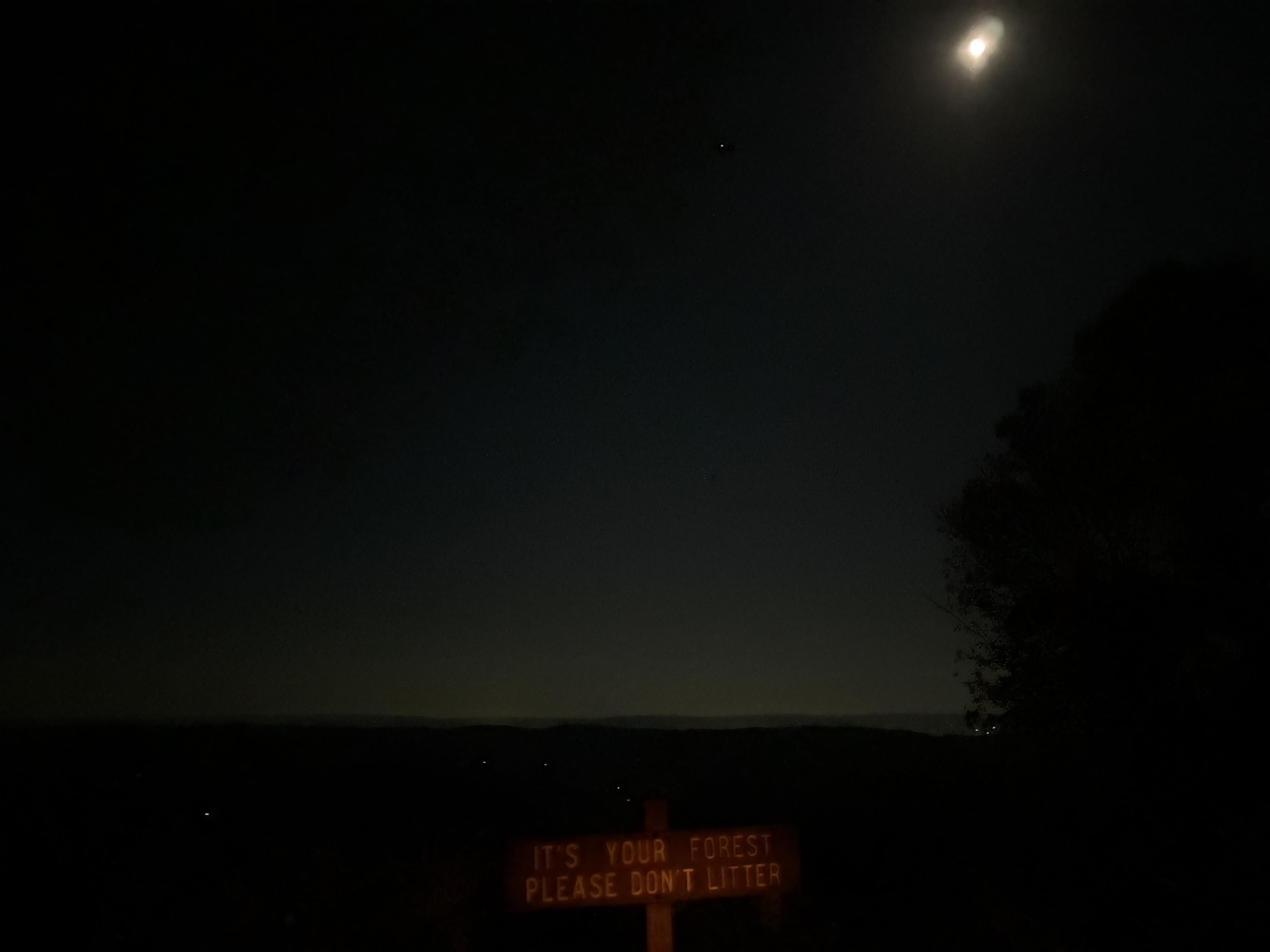 A photo over a valley and the moon at a lookout in Moshannon State Forest in Central PA at night. In the foreground is a sign that reads, “It’s your forest. Please don’t litter.”