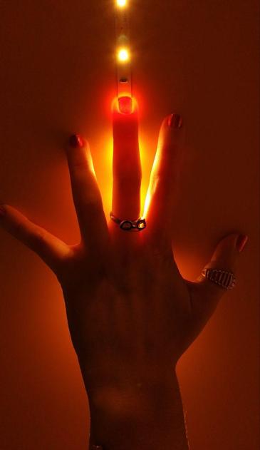Uma mão esquerda, com a palma para cima e os dedos estendidos, é iluminada por uma fita de luzes LED que cria um brilho laranja intenso. O dedo indicador, posicionado diretamente sob a luz, parece brilhar. A mão usa três anéis: um no dedo médio, outro no anelar e um terceiro no polegar. As unhas estão pintadas de vermelho escuro. O fundo é de um tom laranja uniforme.