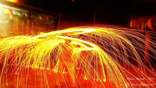 Uma longa exposição de luz cria um espetáculo de faíscas douradas e incandescentes que se espalham como um leque a partir de um ponto central. As faíscas formam arcos e rastros luminosos contra um fundo escuro, com um borrão de luzes e formas desfocadas ao fundo, sugerindo uma estrutura ou paisagem iluminada. A imagem captura um momento dinâmico e energético de fogo em movimento.