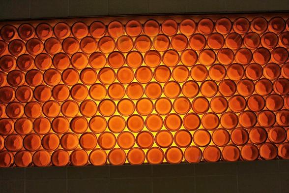 Uma parede é construída com inúmeras garrafas de vidro âmbar, dispostas em fileiras horizontais. A luz brilhante e alaranjada brilha através das garrafas, criando um efeito mosaico luminoso. A parte inferior da imagem mostra uma fileira de azulejos cinzas claros, que contrastam com o brilho quente das garrafas.