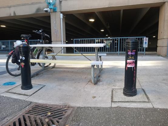 Two black bollards with funny faces painted on them. Behind the bollards is a picnic table with Bob's bike leaning against it, in the background is the UpTown Waterloo parking garage.