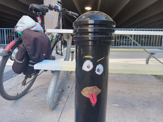 A black metal bollard with a face painted on. The eyes are uneven, with uneven eyebrows, the open mouth is gold-coloured with a red tongue hanging out. It looks vaguely angry, and as if it is blowing a raspberry.

Behind it is a picnic table with Bob's bike leaning on the left, the UpTown Waterloo parking garage is in the background.