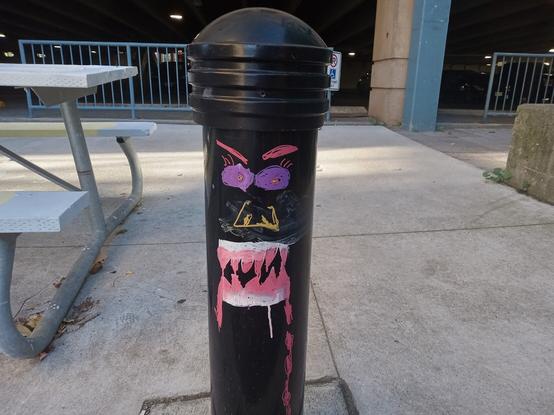 A black bollard with a very angry looking face painted on it. The eyes are purple with three red eyelashes on each eyeball, and red angry eyebrows above. There's squarish yellow nose, and a mouth with white lips and large red teeth. Red drool is coming oyt the right side of the mouth.

The UpTown Waterloo parking garage is in the background.