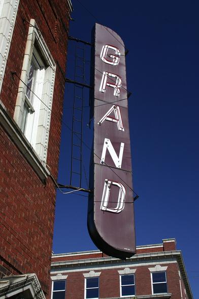 A sign projecting from the corner of the Grand Theater building. It is deep red, with semicircular top and bottom. "GRAND" is displayed vertically in white, italic sans-serif capitals.