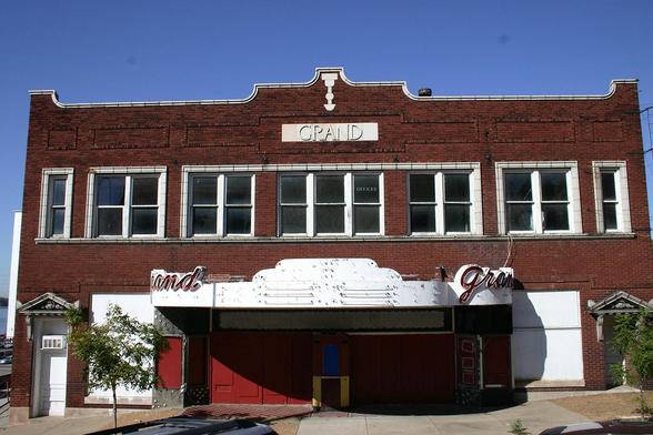 Facade of a vacant movie theater. The box office is centered at the bottom of the frame, its window painted over with blue paint. Above it, an Art Deco-style marquee features neon signs identifying it as the Grand in red script. A bit higher is a row of windows, one of which has serif lettering proclaiming it to be the offices, in all capitals. Above the windows and below an ornate, curved parapet, is a masonry plaque that reads "GRAND" in embossed serif capitals.