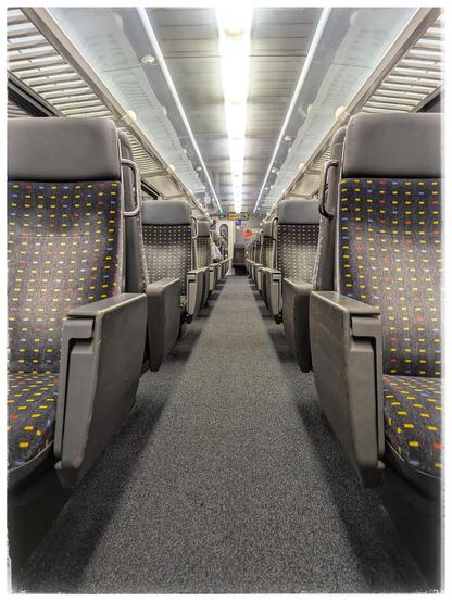 Down to aisle in a railway car, seats can be seen left and right. Only few appear to be taken.