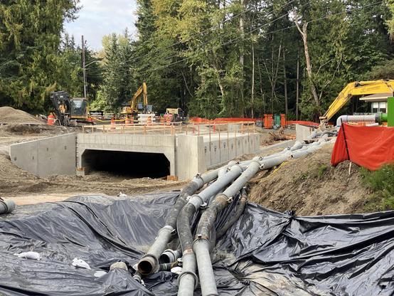 Where a road with a pipe-style water conduit used to be, a salmon-friendly cement culvert which will expose the creek and give it back its width. trees and a cloudy sky are visible beyond; on the right, the arm of a crane; in front/bottom of the image, tarpaulins with temporary piping come towards the viewer.