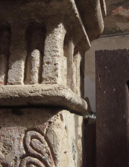 Zwischen einem schönen, alten steinernen Pfosten und der daran befestigten Holztüre ist ein Spalt. Durch diesen beobachtet eine Katze sehr aufmerksam, wer auf der Straße vorbei geht. Man kann nur ein Auge und das Ohr sehen.