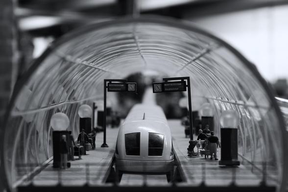 das bild zeigt eine schwarz-weiß-aufnahme einer miniatur-eisenbahnstation unter einer gewölbten struktur, die wie eine überdachung oder ein tunnel aussieht. ein weißer zug steht im mittelpunkt des bildes. entlang des bahnsteigs sind kleine figuren von menschen platziert, die auf den zug warten oder sich mit ihrem gepäck bewegen. schilder mit der aufschrift "hauptbahnhof" sind an den pfosten angebracht und zeigen die zeit an. der fokus des bildes ist relativ scharf auf den zug und die figuren, während der hintergrund und die überdachung leicht verschwommen sind.