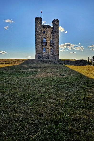 A low-angle, sunlit photograph of Broadway Tower, a tall, gothic-style stone folly with two prominent round turrets, standing atop a grassy hill. The sun is setting behind the tower, casting long shadows across the dry, golden-brown grass in the foreground and creating a dark silhouette around the structure against a bright blue sky with scattered white clouds. A small flag is visible on the roof.