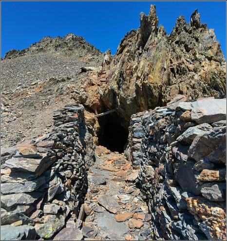 Une entrée en cavage pour une mine.
De chaque côté des murets en pierres sèches protège l'accès.
À l'arrière plan, les crètes d'une montagne sous un ciel bleu.