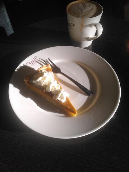 Auf einem dunkelbraunen Holztisch steht ein von der Nachmittagssonne angeleuchteter weißer Teller mit einem Stück Pumpkin Pie darauf. Dabei handelt es sich um einen Kuchen, der aus zwei Schichten besteht: Eine cremige, würzige, süße Kürbis-Eier-Sahne-Crème bettet sich auf buttrigen Mürbeteig. 
Auf dem Kuchenstück thronen drei Tuffs aus frischer Schlagsahne. 
Dahinter steht ein Becher mit heißem Cappuccino mit Karamell.