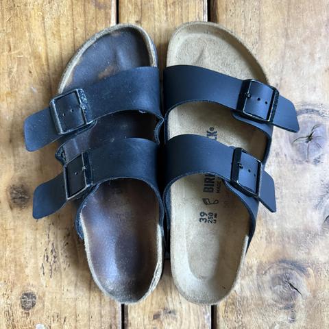 A pair of black sandals with two straps and buckles, placed on a wooden surface. One sandal appears worn, with a visible scuff on the sole.