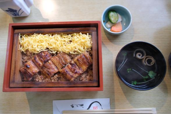 Barbecued eel slices and shredded cooked eggs over seasoned rice; pickles on top right; a bowl of clear broth soup, and chopsticks at the bottom