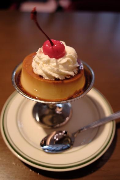 A cudtard/flan on a silver stand with white whipped cream and a maraschino cherry on top