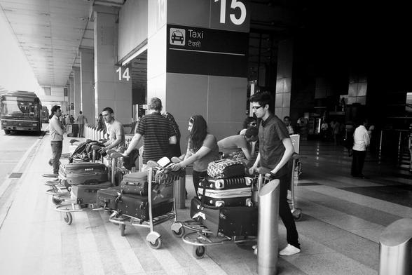 Schwarzweißfoto, aufgenommen an einem Flugplatz in Indien. Eine Reihe von Leuten mit Koffern auf Trolleys wartet darauf, in den nahenden Bus einzusteigen.
