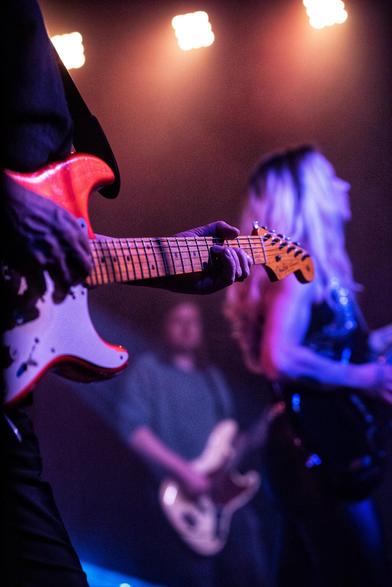 Der Focus liegt auf dem Griffbrett einer E Gitarre. Dahinter erkennt man verschwommen noch andere Musiker.