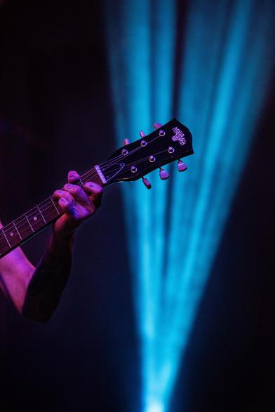 Im Vordergrund ein Gitarrenhals der von tätowierten Fingern bespielt wird. Im Hintergrund ein Lichtstrahl eines Bühnenscheinwerfer.