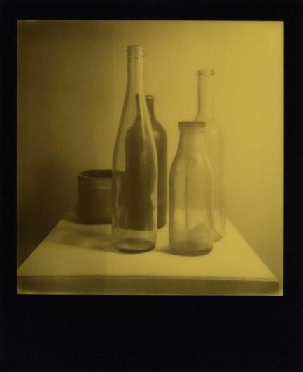 Ein Stilleben mit drei durchsichtigen Glasflschen im Vordergrund, einer Tonflasche und einem Tongefäß im Hintergund. Alles steht auf einm weißen Sockel vor heller Wand. Das Foto ist in Gelb- und Grautönen gehalten, weil es ein Schwarz-gelb-Film von Polaroid ist, mit einem schwarzen Bildrahmen.