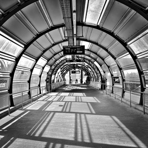 A view along a circular (above ground) tunnel with a sign advertising an upcoming train. Morning shadows cross the floor from the windows on the left side.