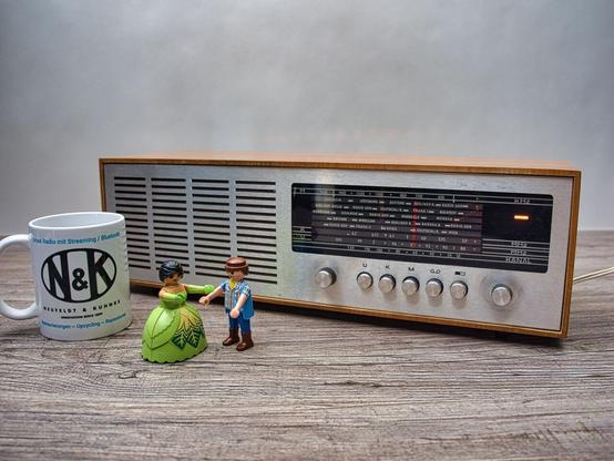 Ein Tischradio mit gebürsteter Aluminium Front, eingeschaltet.
Die Bereitschaftsleuchte ist oben rechts zzu erkennen , sie leuchtet in beige.
Vor dem Radio stehen 2 Playmobil Figuren. Links neben den Figuren steht ein N&K Kaffeebecher.
Das Radio hat keine Skalenbeleuchtung.
Knöpfe für Lautstärke, U K M , Tonband und Stereo, sowie Sendereinstellung sind ebenfalls in Alu poliert ausgeführt und befinden sich unter dem Skalenband.
