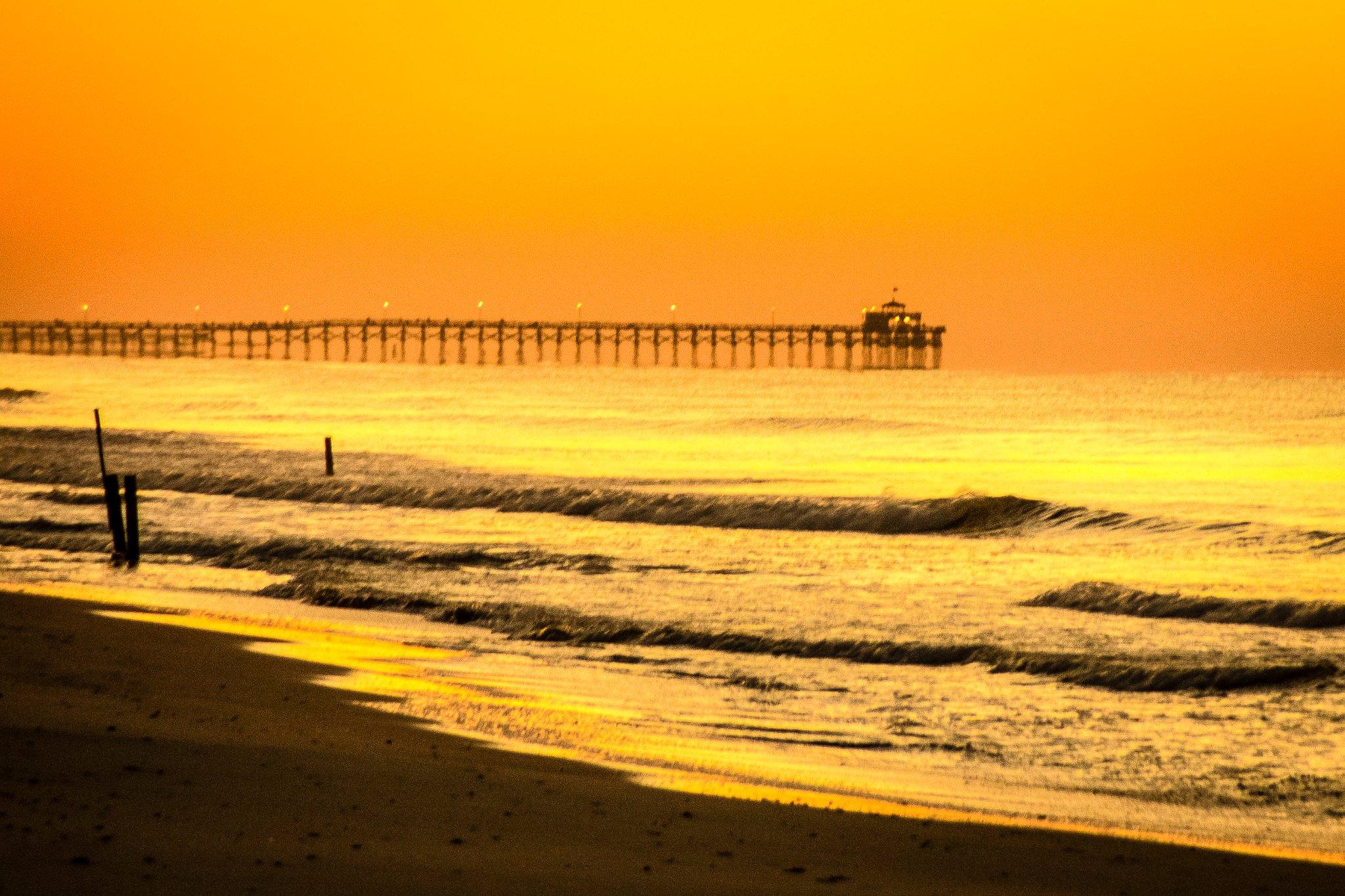 A golden sky and ver the ocean as waves hit the beach with a fishing pier in the background.