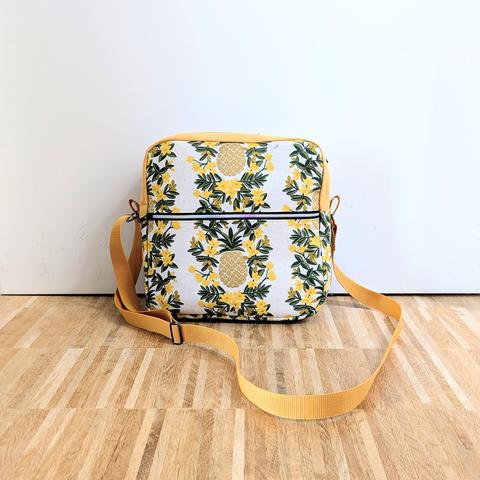 Handbag with a flower-pineapple pattern, the pineapples are golden. It has ha square pattern with a zipper on top and roughly at golden cut in the front.