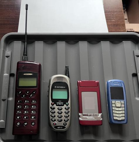 A collection of four vintage mobile phones, displayed on a gray tray. From left to right: a dark wood coloured Benephon with a long antenna, a silver Motorola model, a shiny red Nokia flip phone, and a blue Nokia phone with a keypad.